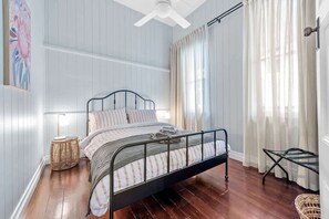 Un dormitorio con estructura de cama metálica, lámpara de noche, cesta de mimbre y ventana con cortinas.