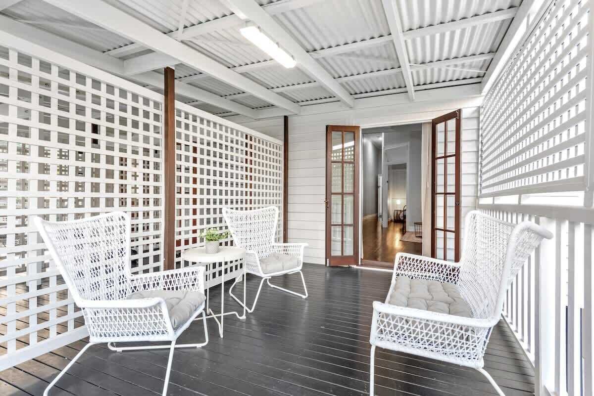 Un espacio exterior techado con mobiliario de mimbre blanco, una mesa de madera con una planta y vistas a una habitación contigua a través de una puerta de vidrio.