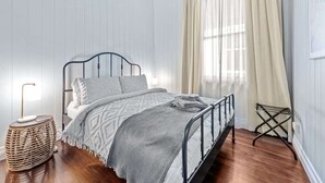 A neatly arranged bedroom with a metal bed frame, a bedside lamp, a woven side table, and a window with curtains. - The Deck! 2 Bed/1 Bath/1 Car ~ New Farm (Brisbane)