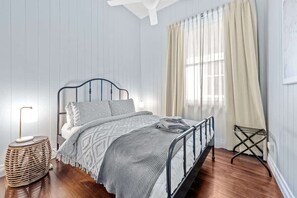 Ein ordentlich eingerichtetes Schlafzimmer mit einem Metallbettgestell, einer Nachttischlampe, einem gewobenen Beistelltisch und einem Fenster mit Vorhängen.