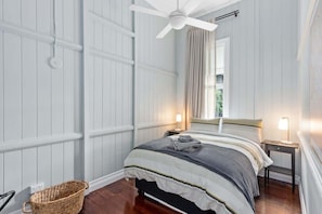 Ein Schlafzimmer mit einem Bett, einem Deckenventilator, einem Nachttisch, einer Lampe und einem Korbwäschesammler.