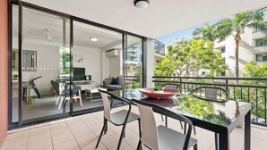 Ein moderner Balkon mit Glastisch und -stühlen, einem Essbereich und Blick auf ein Wohngebäude und Grünflächen.