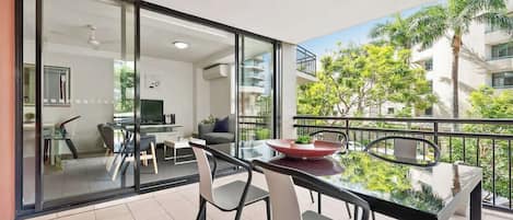 A modern balcony with a glass table and chairs, a dining area, and a view of a residential building and greenery.