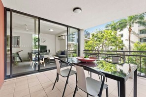 Ein moderner Balkon mit Glastisch und -stühlen, einem Essbereich und Blick auf ein Wohngebäude und Grünflächen.