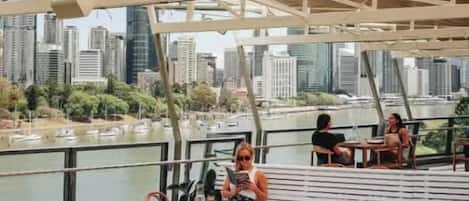 A dining area with tables and chairs, overlooking a body of water and a city skyline.