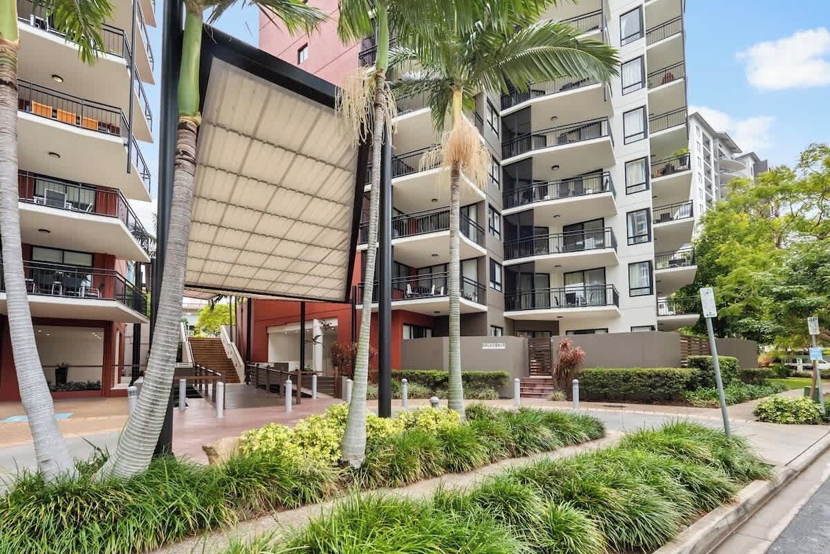 A multi-story residential building with balconies, a covered outdoor area, and palm trees.