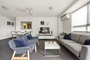 Ein modernes Wohnzimmer mit einer Couch, Sesseln, einem Couchtisch, einem Fernseher und einem Deckenventilator.