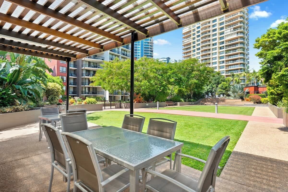An outdoor patio with a glass table and chairs, surrounded by greenery and a view of high-rise buildings.