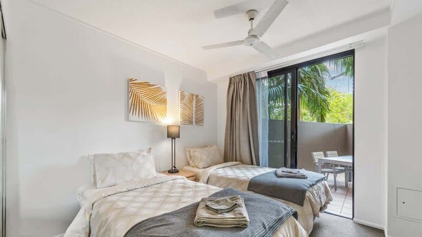 Ein Hotelzimmer mit zwei Betten, einem Deckenventilator, einem großen Fenster mit Vorhängen und Blick auf Grün.
