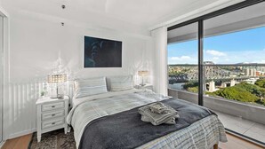 Ein Schlafzimmer mit einem großen Bett, einem Nachttisch, einer Lampe und Blick auf eine Stadtlandschaft.