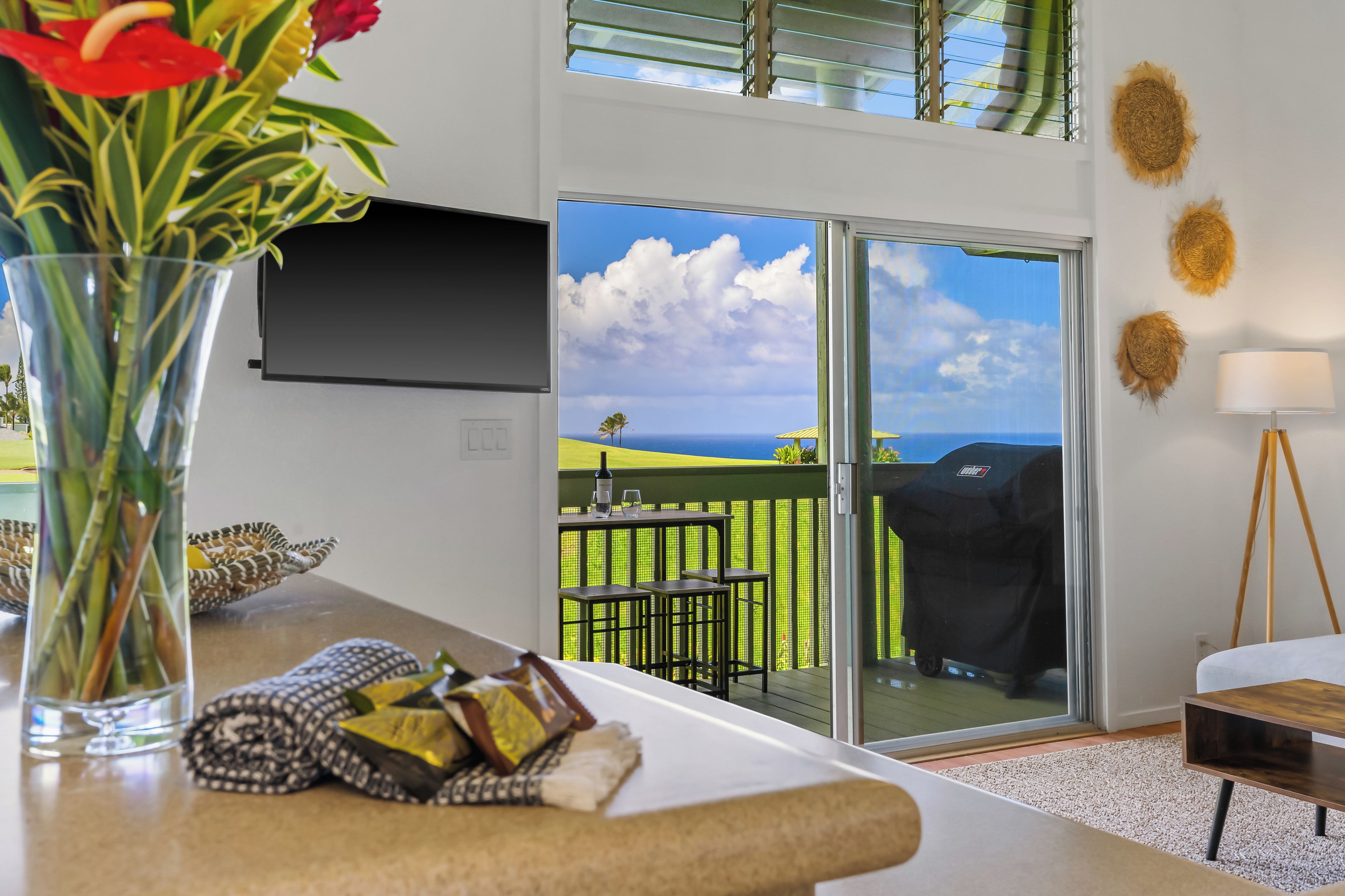 A modern living room with a large window, a flat-screen TV mounted on the wall, and a dining area with outdoor seating.