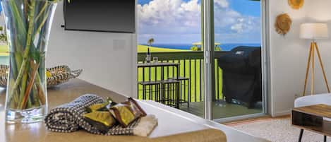 A modern living room with a large window, a flat-screen TV mounted on the wall, and a dining area with outdoor seating.