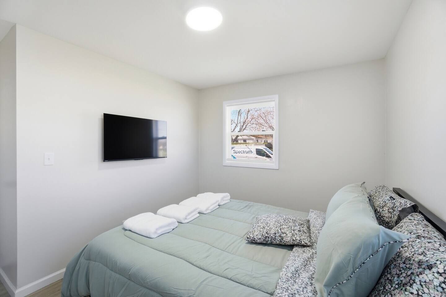 A bedroom with a bed, a flat-screen TV, a window with a view, and a wall-mounted light.