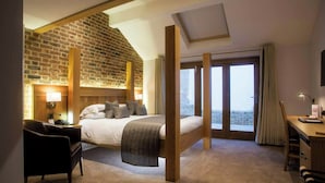 A hotel room with a brick wall, a bed with a wooden headboard, a desk, a chair, and a balcony with curtains. - Tewin Bury Farm Hotel (Welwyn)