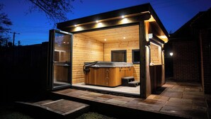 A hot tub with a wooden exterior and glass doors, set in a backyard with a brick wall and a wooden deck. - Tewin Bury Farm Hotel (Welwyn)