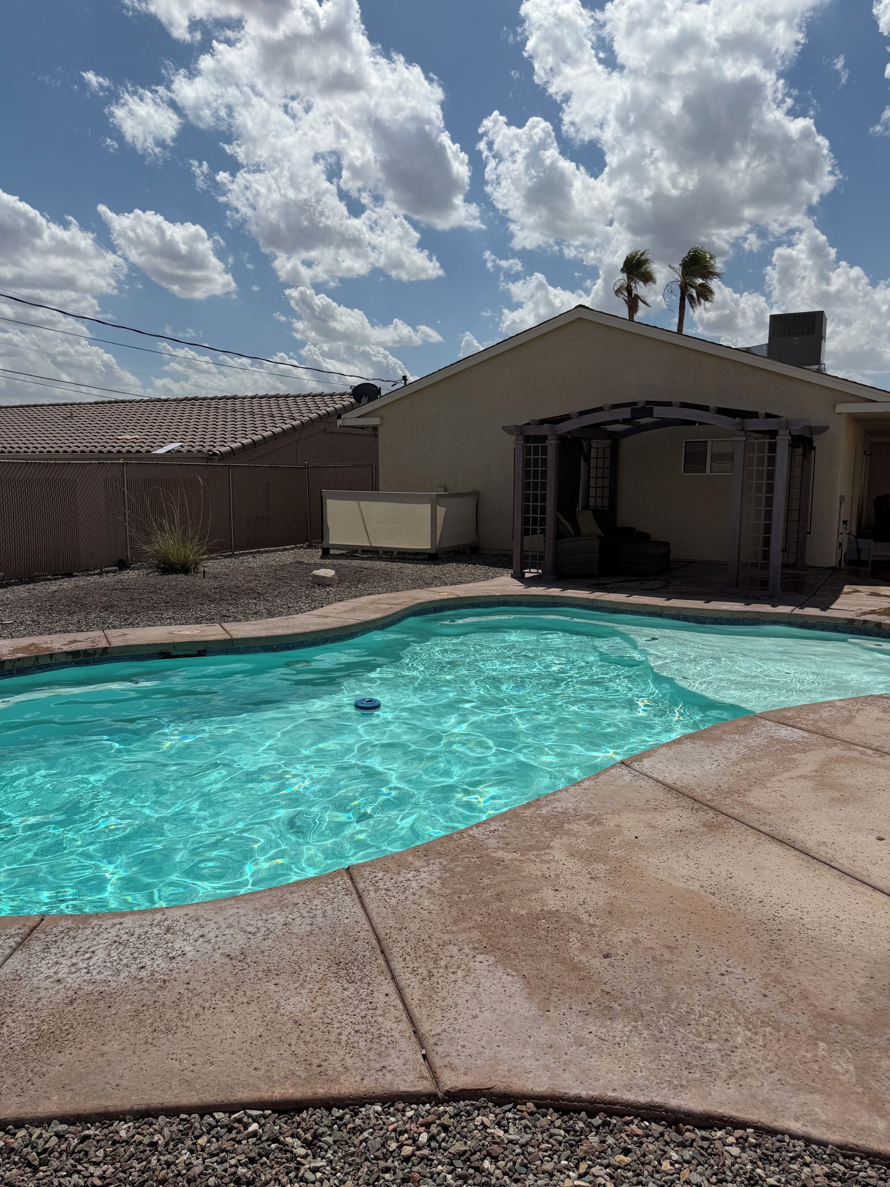 En swimmingpool med en klar blå vandoverflade, omgivet af en betonflisebelagt terrasse. Der er en overdækket patio med en pergola og en mindre bygning i baggrunden.