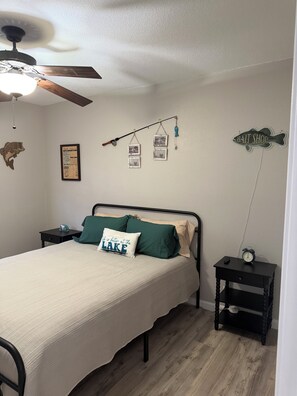 Ein Schlafzimmer mit einem Bett, einem Nachttisch, einer Wanduhr, einem Deckenventilator und Wanddekorationen, darunter ein Schild mit einem Fischmotiv und gerahmte Fotos.