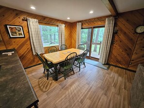 A dining area with a wooden table and chairs, a clock on the wall, and large windows with curtains. - Updated cabin on the Au Sable River 10 minutes from Grayling (Grayling)