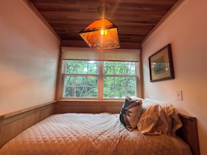 A bedroom with a large window, a quilted bed, a hanging light fixture, and a framed picture on the wall.