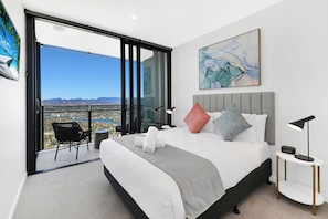A modern bedroom with a large bed, a view of a cityscape through a sliding glass door, a small table with a lamp, and a painting on the wall. - Stunning Water View 1BR  | Steps to Casino and Tram (Broadbeach)