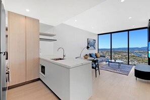 Una cuina moderna amb una illa central, una zona de menjador amb un sofà i taula, i un gran finestral que ofereix vista a la ciutat.