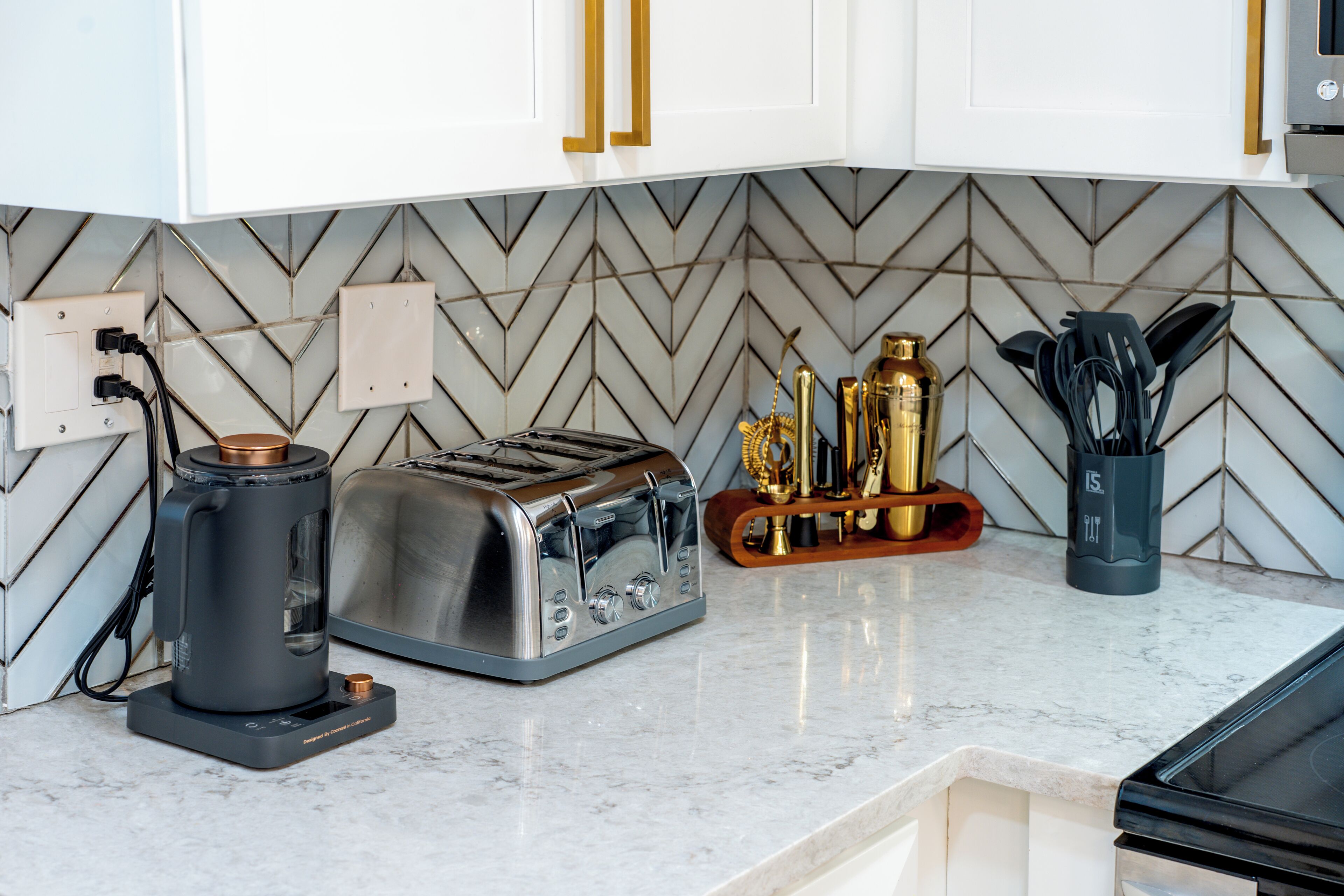 A kitchen with a modern design, featuring a herringbone tile backsplash, a toaster, a coffee maker, and a utensil holder.