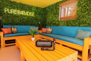 Ein Zimmer mit grüner Wand, einem neonbeschrifteten Schild mit 'PURE PARADISE', einem blauen Sofa und einem Holztisch mit Lautsprecher und Mikrofon.