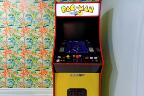 Ein Pac-Man-Spielautomat mit gelbem Unterteil und rotem Rahmen, vor einer Wand mit tropischem Wandpapier platziert.