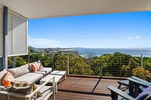 Ein Balkon mit Holzboden, ein weißes Sofa mit gestreiften Kissen und Blick auf das Meer und die Hügel.