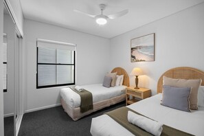Hotelkamer met twee bedden, een plafondventilator, een raam met zonwering, een nachtkastje met een lamp en een schilderij aan de muur.