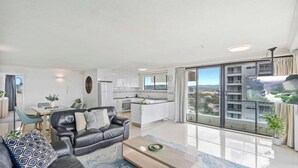 A modern living room with a sofa, coffee table, and dining area. A kitchen is visible in the background, and the room has large windows with a view of a cityscape. - Water Views at Pacific Keys Central - Hosted By Gold Coast Holiday Stays (Mermaid Waters)