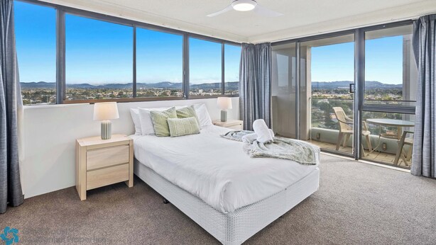 A hotel room with a large bed, a nightstand, a lamp, and a view of the city through large windows. - Water Views at Pacific Keys Central - Hosted By Gold Coast Holiday Stays (Mermaid Waters)