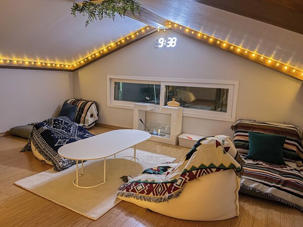 A cozy loft bedroom with a sloped ceiling, a small table, bean bag chairs, a window, and a clock displaying 9:38.
