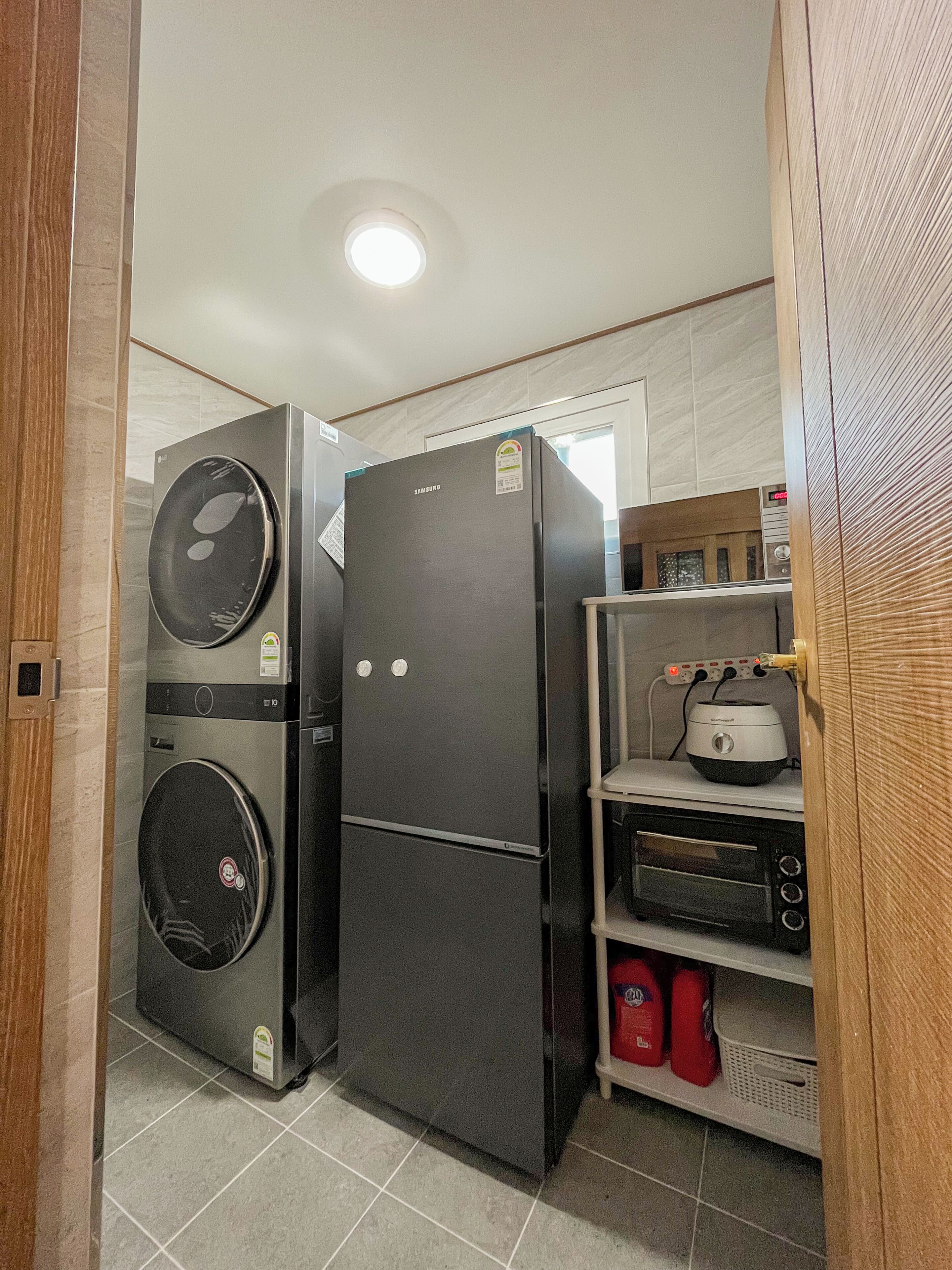 Una cocina compacta con un refrigerador Samsung, lavadora, tostadora y un estante con productos de limpieza.