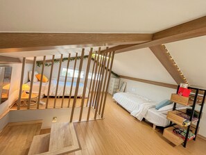 A loft bedroom with a sloped ceiling, wooden beams, a bed with white bedding, a small table, and a shelf with books.