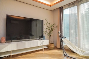 A modern living room with a flat-screen TV, a small plant, a hammock, and a large window with sheer curtains. - Cozy Family House in the nature of Ganghwa Island with BBQ & Campfire (Ganghwa-gun)