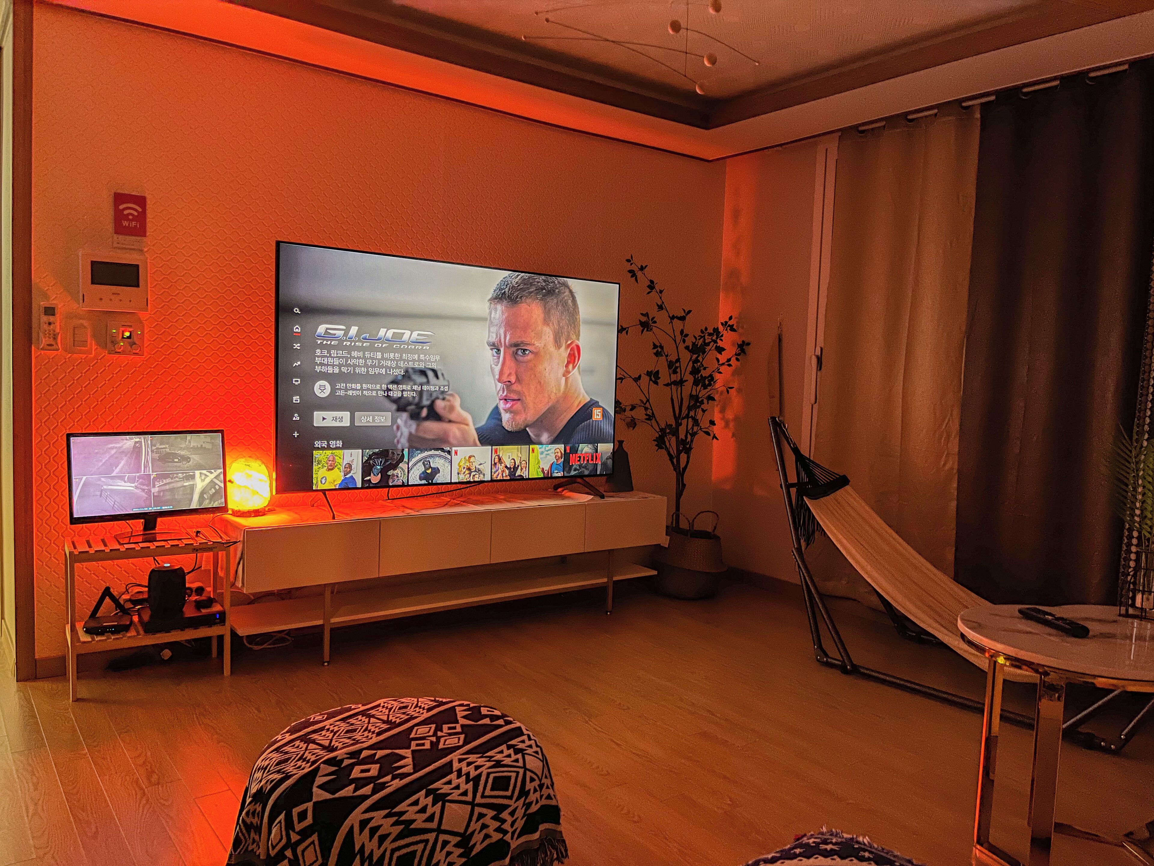 A living room with a large flat-screen TV mounted on the wall, a smaller monitor on a stand, a plant, and a foldable chair.