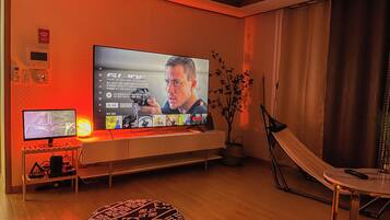 A living room with a large flat-screen TV mounted on the wall, a smaller monitor on a stand, a plant, and a foldable chair.