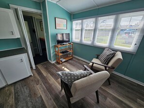 A room with teal walls, a wooden entertainment center, a television, a sofa, and a window with blinds. - Lyons Cottages - #7 King Studio Walk to the Beaches of Stanhope National Park! (Stanhope)