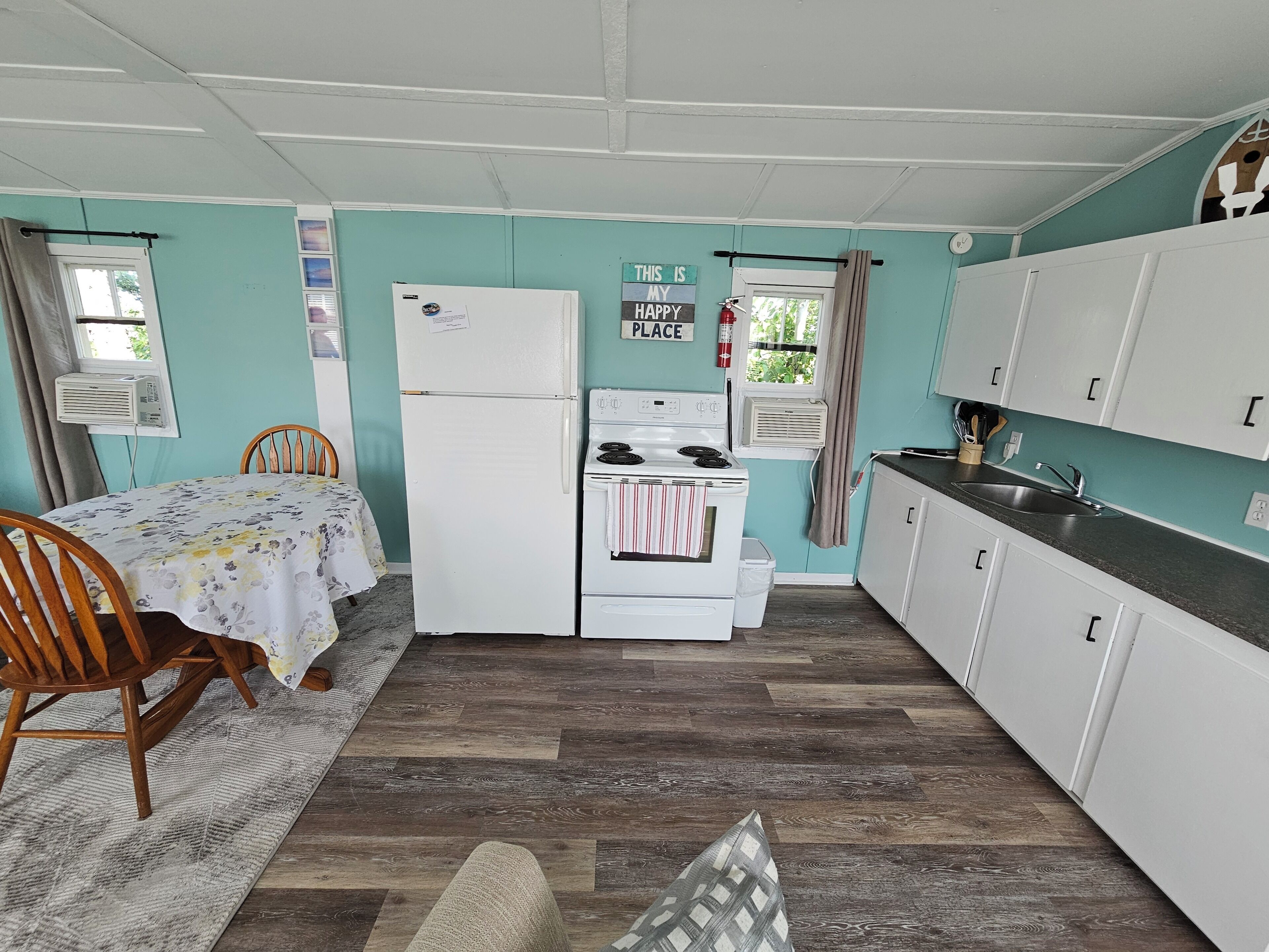 A compact kitchen with white cabinets, a refrigerator, a stove, and a sink. There is a dining table with chairs and a rug on the floor.