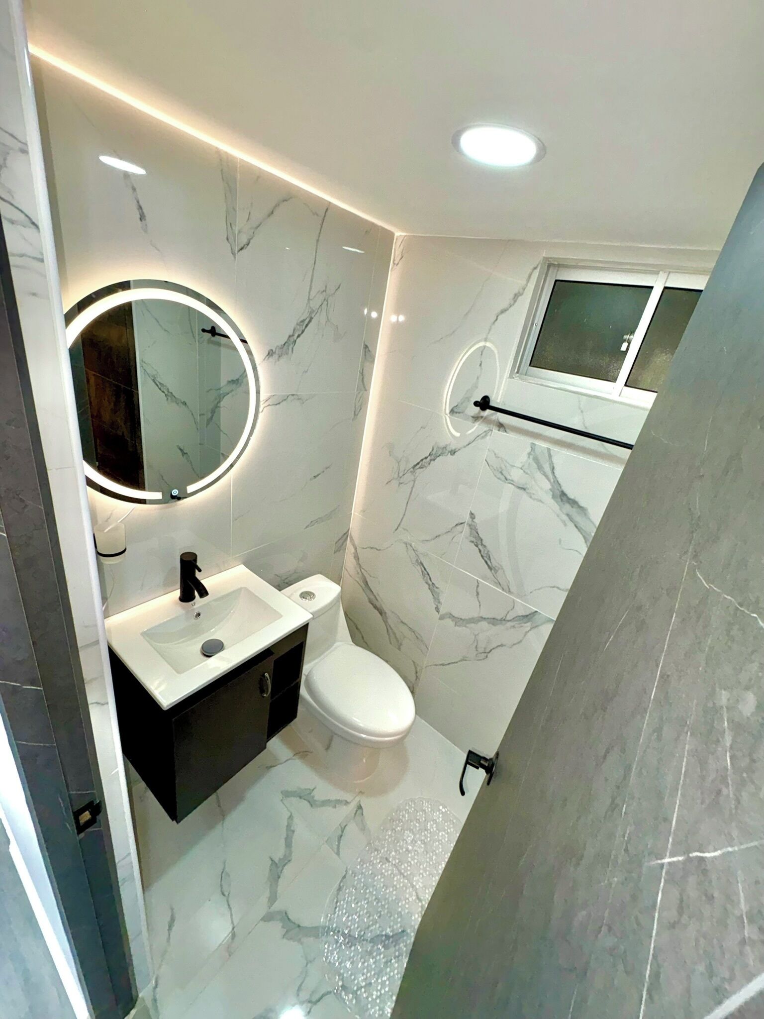 Ein modernes Badezimmer mit Marmorboden, einem runden Spiegel mit Beleuchtung, einer weißen Toilette und einem schwarzen Schrank.