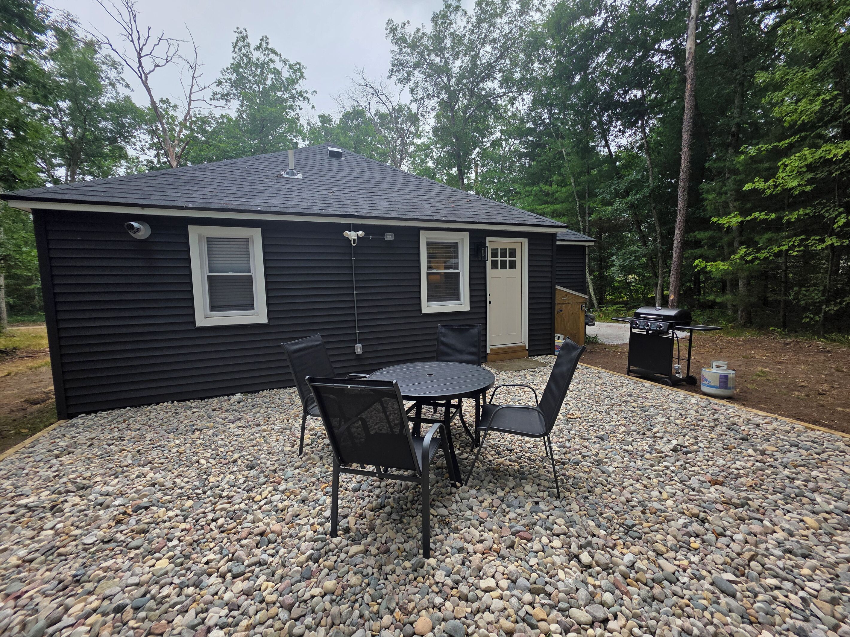 A small house with a patio area featuring a table and chairs, surrounded by a gravel driveway, and a grill in the background.