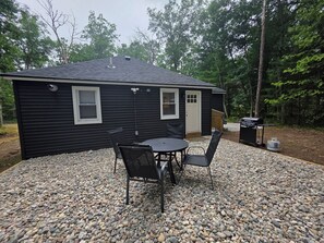 A small house with a patio area featuring a table and chairs, surrounded by a gravel driveway, and a grill in the background. - The Haven at Idlewild (Idlewild)