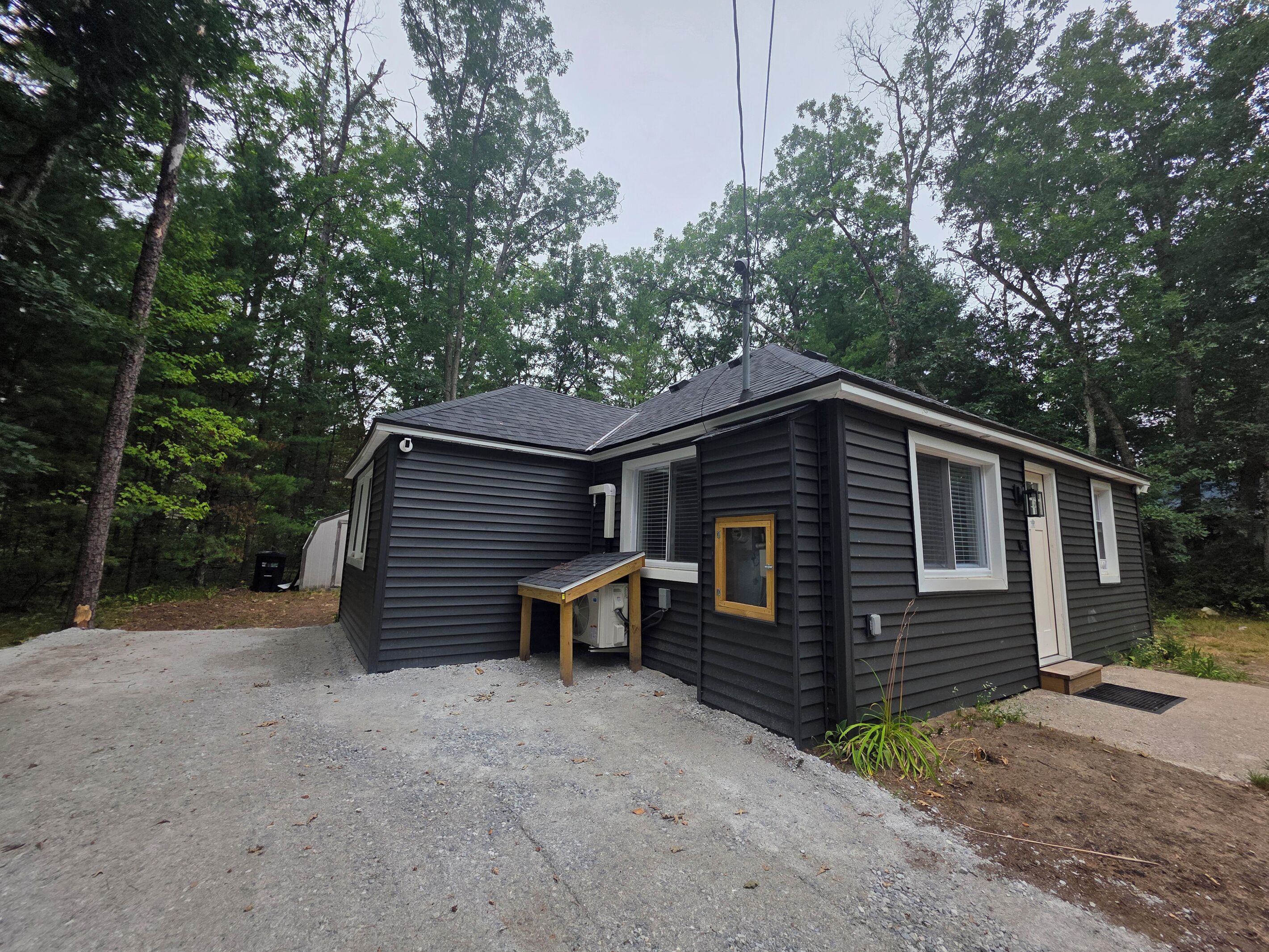 A small, dark-colored cabin with a gravel driveway, surrounded by trees.