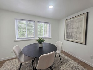 A dining area with a round table, four chairs, a potted plant, and a large abstract painting on the wall.