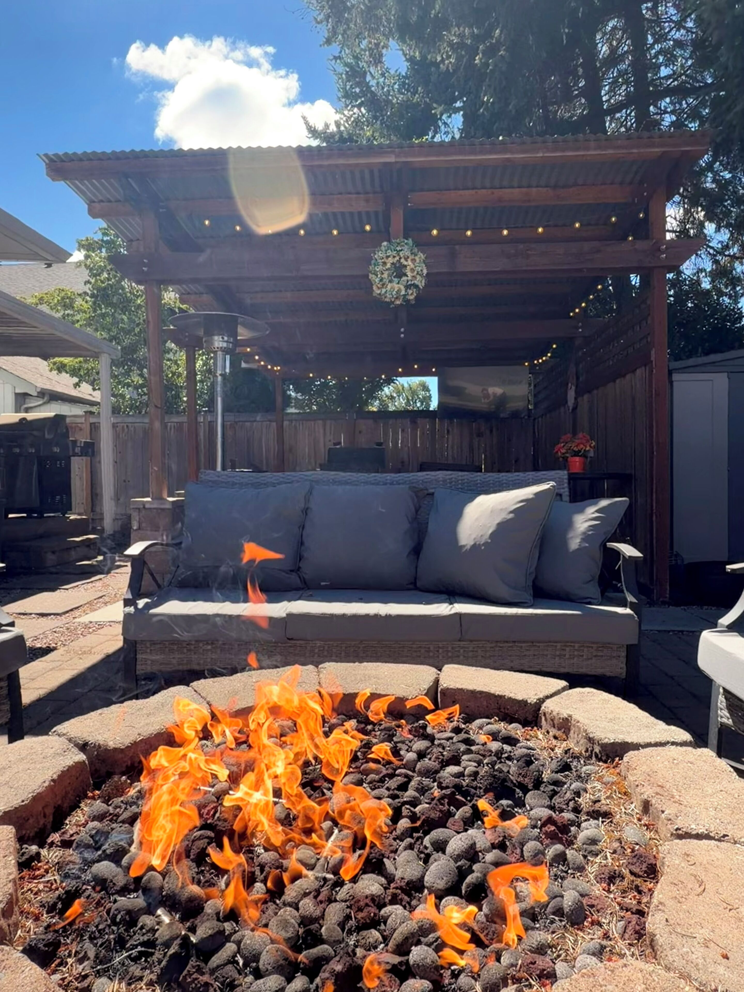 A fire pit with a wooden pergola overhead, a cushioned outdoor seating area, and a clear sky.