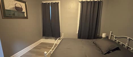 A bedroom with a single bed, two windows with curtains, a framed picture on the wall, and a white headboard.