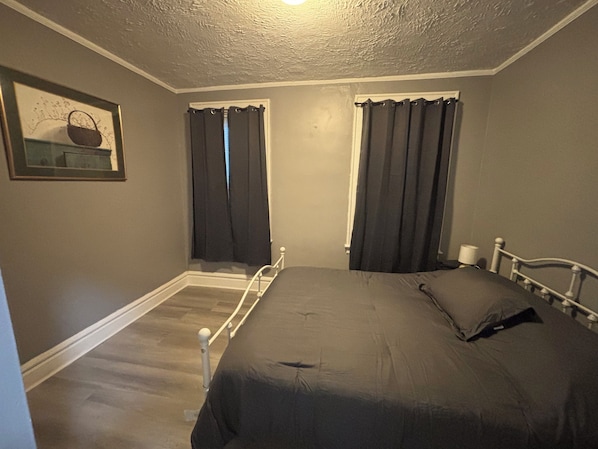 A bedroom with a single bed, two windows with curtains, a framed picture on the wall, and a white headboard.