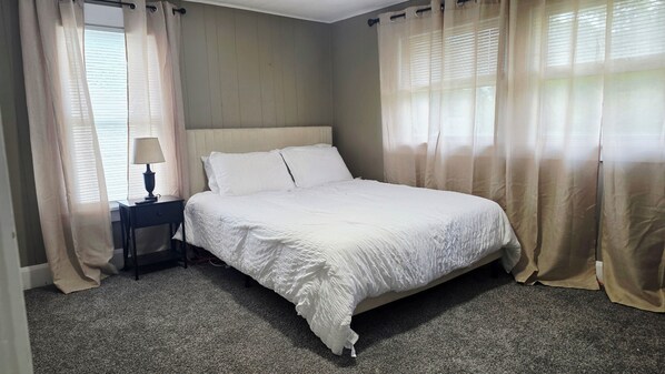 A neatly made bed with white bedding in a room with a carpeted floor and a nightstand with a lamp.