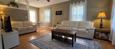 A living room with a sofa, a coffee table, a TV on a stand, a ceiling fan, and a welcome sign.